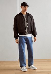 Giubbotto bomber nero con bottoni a pressione argentati, maglietta grigia, jeans blu relaxed fit e sneakers bianche con dettagli grigi, completato con un cappellino nero.