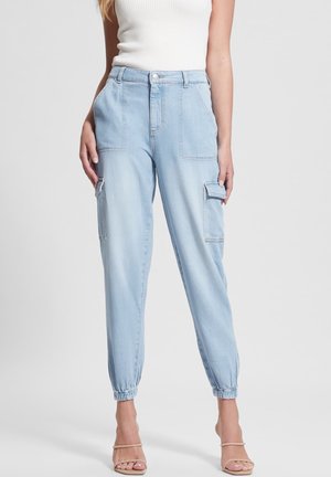 Femme portant un pantalon cargo en denim bleu clair taille haute avec des poignets élastiques et des poches plaquées à l'avant et sur les côtés, associé à un haut blanc sans manches.
