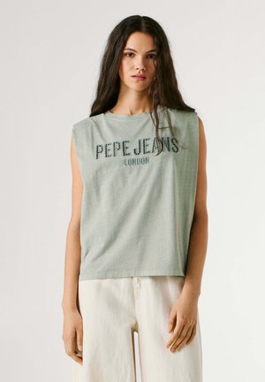 Jeune femme aux longs cheveux noirs portant un haut sans manches vert clair avec le texte « Pepe Jeans London » et un pantalon blanc, sur un fond uni.