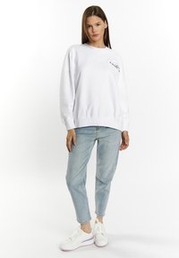 Felpa bianca con scollo tondo e vestibilità comoda, caratterizzata da un design di nuvola ricamato sul petto, abbinata a jeans azzurri chiari e sneaker bianche.