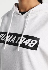 Šedá mikina s kapucí a černým grafickým páskem s nápisem "PUMA 1948." Obsahuje stahovací kapuci a měkkou texturu tkaniny.