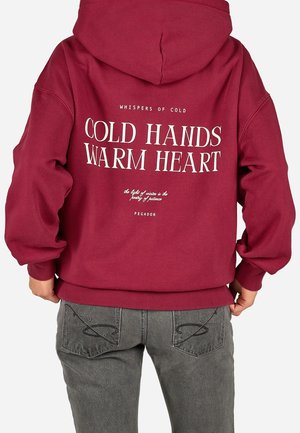 Osoba nosząca czerwoną bluzę z kapturem z napisem "Cold Hands Warm Heart" na plecach, w połączeniu z szarymi dżinsami z naszywanymi wzorami na kieszeniach.