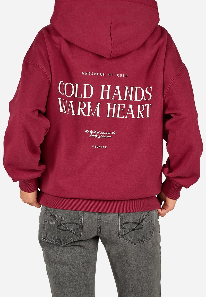 Personne portant un sweat à capuche rouge avec le texte "Cold Hands Warm Heart" dans le dos, associé à un jean gris avec des poches décorées de coutures.