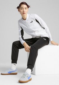 Puma trenirka koja se sastoji od bijele jakne s patentnim zatvaračem i crnim detaljima te crnih suženih hlača. Model nosi sive i plave tenisice, preko bijelih čarapa.
