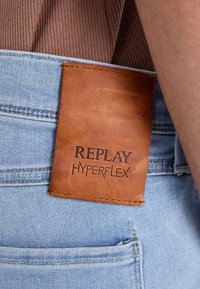Los jeans de mezclilla azul claro presentan un parche de cuero marrón con el texto en relieve "REPLAY HYPERFLEX" en la cintura trasera.