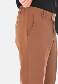 Primo piano di una persona che regola la vita di un pantalone marrone con passanti per cintura, tasche laterali e una tasca posteriore con bottone.