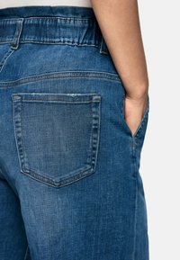 Denim jeans with a medium blue wash, featuring a back pocket, subtle whiskering, and a relaxed fit with an elastic waistband.