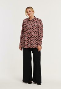 Mehrfarbiges Hemd mit Knöpfen auf schwarzem Grund, das ein geometrisches Muster in Rot und Creme aufweist. Kombiniert mit weiten schwarzen Hosen.