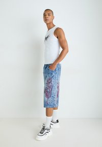 Weißes Tanktop mit grafischem Design, dekorative blaue Jeansshorts mit rot-weißen Mustern und schwarz-weiße Sneaker mit dicken Sohlen.