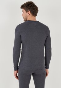 Maglietta grigia a maniche lunghe con scollo rotondo, design aderente e polsini a costine. Il tessuto appare morbido e leggermente testurizzato, adatto per essere indossato a strati.