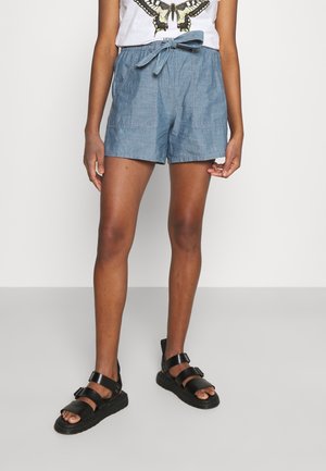 Personne portant un short bleu à nouer à la taille, un t-shirt blanc avec un imprimé papillon et des sandales noires à boucle, debout sur un sol gris.