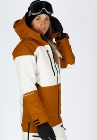 Giacca da neve arancione e bianca; presenta un cappuccio, tasche con zip e cordini regolabili; tessuto testurizzato; abbinata a guanti neri.