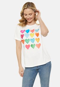 Hvid kortærmet t-shirt med en farverig hjerte-graphic i forskellige nuancer, herunder pink, blå, grøn og orange. Denim jeans iført.