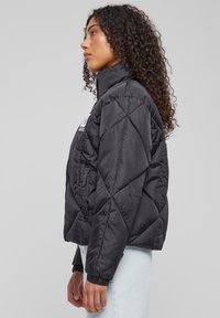 Juodasis pūkuotas pufferis su aukštu apykakle, elastinėmis rankovėmis ir logotipo detale. Turi trumpą dizainą su lygiu, blizgiu paviršiumi.