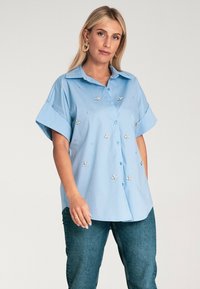 Jasnoniebieska koszula z krótkim rękawem i podwiniętymi rękawami, ozdobiona haftem w kwiaty i drobnymi detalami. Wykonana z miękkiego materiału i o luźnym kroju.