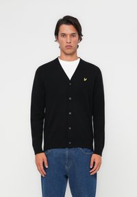 Black cardigan with a V-neck, front buttons, and a small yellow logo on the left chest. Worn over a white shirt and blue jeans.