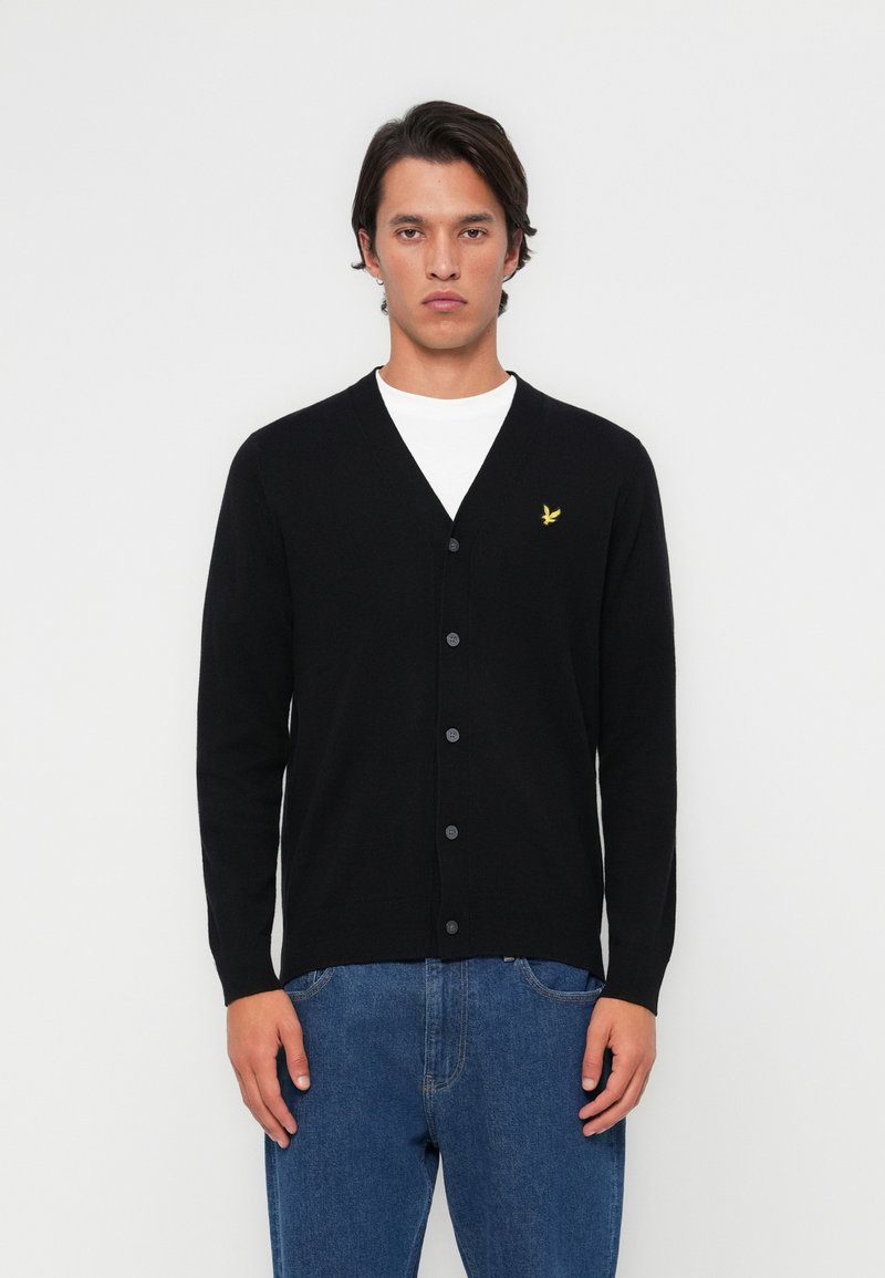 Black cardigan with a V-neck, front buttons, and a small yellow logo on the left chest. Worn over a white shirt and blue jeans.