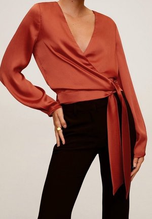 Femme portant un chemisier cache-cœur en satin couleur rouille à manches longues et un pantalon noir, posant avec une main sur la hanche et l'autre détendue.