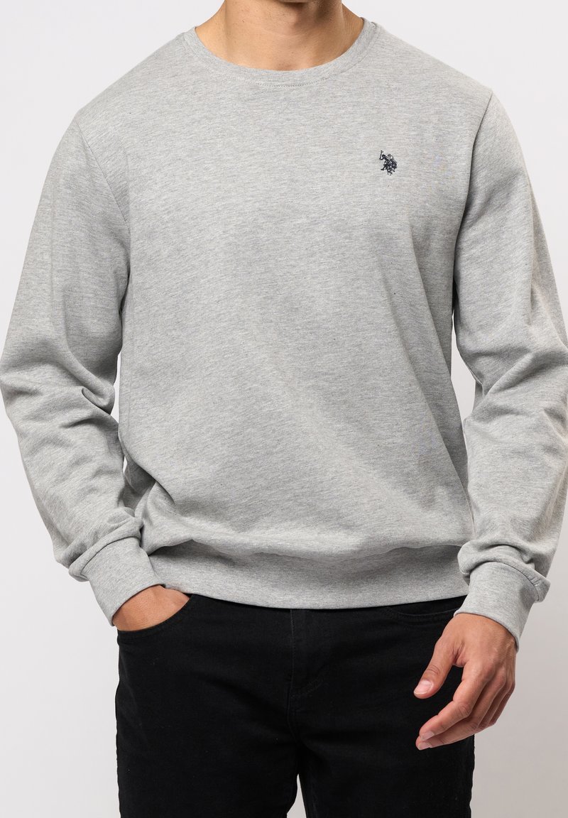 Homme portant un sweat-shirt à manches longues gris clair avec un petit logo sombre sur le côté gauche de la poitrine et un pantalon noir, mains détendues, fond neutre.
