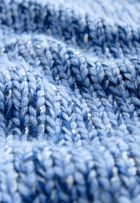 Tekstūrēta adīta auduma virsma dažādos zilo toņos, ar mīkstu, pūkainu izskatu un periodiski sastopamiem baltajiem plankumiem.