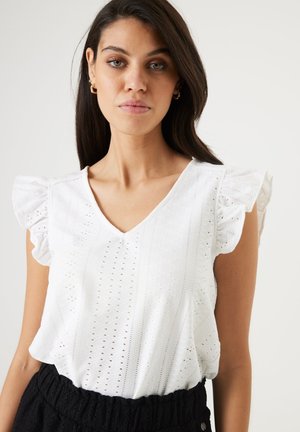 Blusa blanca de manga corta con escote en V y acentos con volantes, con detalles de ojales en una tela ligera. Falda negra texturizada abajo.