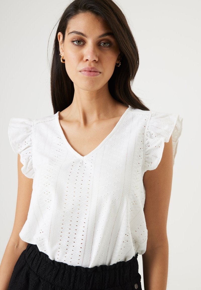 Blusa blanca de manga corta con escote en V y acentos con volantes, con detalles de ojales en una tela ligera. Falda negra texturizada abajo.