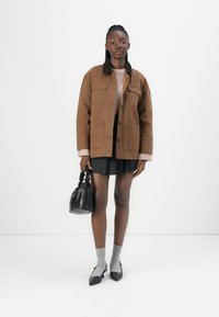 Jachetă oversized maronie cu buzunare aplicate, guler negru, pulover roz, fustă mini neagră, șosete gri și pantofi cu toc punti negru. Geantă de mână neagră.