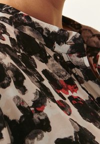 Blusa stampata floreale con colletto, caratterizzata da un mix di motivi astratti in nero, grigio e rosso su uno sfondo chiaro. Tessuto morbido.