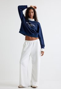 Sudadera corta azul marino con texto blanco, combinada con pantalones blancos de corte holgado. El conjunto presenta puños acanalados y una textura de tela suave.