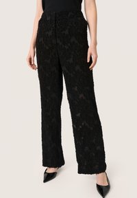 Pantalones anchos negros de tela texturada con dobladillo deshilachado. El material tiene una apariencia de capas y pelusa. Combinados con zapatos negros de punta.