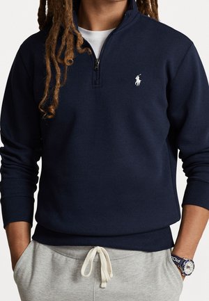 Persona vistiendo un sudadera de cremallera corta azul marino con el logo de polo blanco, pantalones deportivos gris claro con cordón y un reloj de pulsera en la muñeca izquierda.