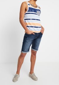 Pruhovaný top bez rukávů v modré, bílé a broskvové barvě, s kapsičkou na prsou; v kombinaci s tmavými džínovými šortkami a světlými šedými lodními botami.