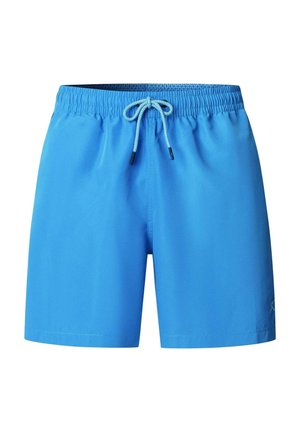 Hombre musculoso de pie, con pantalones cortos de baño azul brillante con cordón, zapatos beige sin cordones, expresión neutral, fondo gris claro.