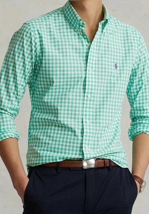 Mężczyzna ubrany w zielono-białą kraciastą koszulę zapinaną na guziki z podwiniętymi rękawami, brązowy pasek, granatowe spodnie i zegarek na nadgarstku.