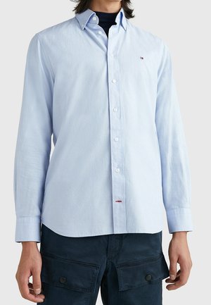 Jasnoniebieska koszula zapinana na guziki noszona na czarnym podkoszulku, zestawiona z ciemnoniebieskimi spodniami typu cargo z klapkami na kieszeniach.