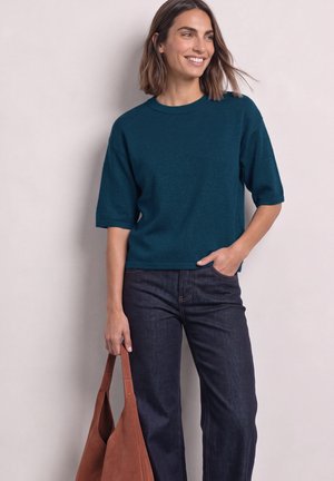 Femme aux cheveux bruns mi-longs portant un pull à manches courtes bleu sarcelle et un jean foncé, tenant un sac cabas en cuir marron.