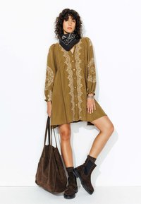 Robe chemise vert olive avec des broderies blanches intricées, assortie à un sac fourre-tout en suède marron et des bottines marron avec des accents noirs.