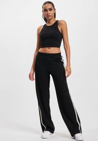 Kvinne som står med hendene avslappet, iført et svart ermeløst croptop, svarte vide bukser med hvite sidestriper, og hvite joggesko.