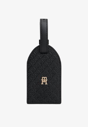 Fekete bőr poggyászcímke dombornyomott mintával, lekerekített tetejével és aranyszínű logóval a közepén. Masszív rögzítőhurokkal rendelkezik.