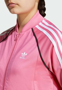 Ružová Adidas tréningová bunda s predným zipsom, rebrovaným golierom a tromi bielymi pruhmi na rukávoch s čiernymi akcentmi.