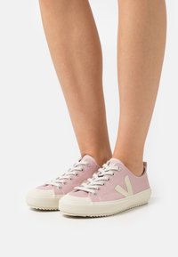 Baskets en toile rose clair avec semelles en caoutchouc blanches, présentant des accents et des lacets couleur crème, et un logo « V » sur chaque côté.