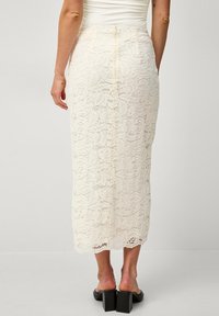Jupe midi en dentelle crème avec un motif floral, présentant une coupe droite et une fermeture éclair au dos. Texture lisse avec un design délicat.