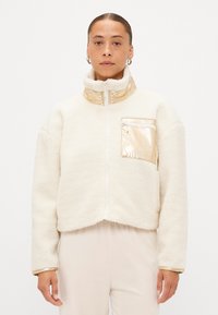 Jachetă albă din fleece cu guler înalt, accente aurii pe guler și buzunar, și un design scurt, având un fermoar frontal.