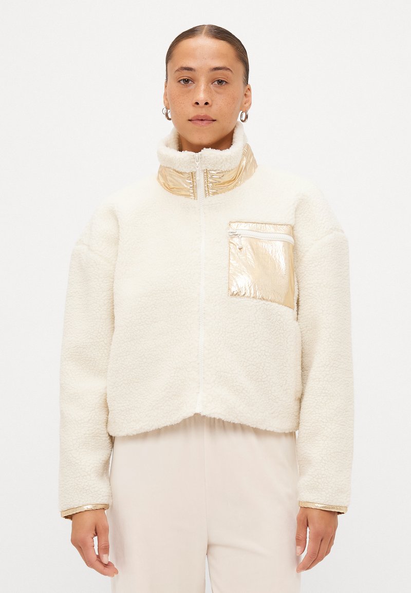 Jachetă albă din fleece cu guler înalt, accente aurii pe guler și buzunar, și un design scurt, având un fermoar frontal.
