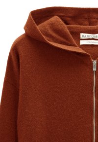Kardigan z kaszmiru w kolorze rdza, z złożonym kapturem, przednim zamkiem błyskawicznym oraz gładką, miękką fakturą. Metka wskazuje na 100% materiał z kaszmirem.
