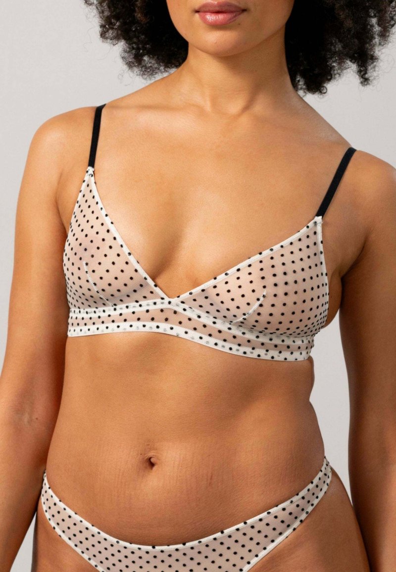 Bralette beige transparent avec des pois noirs, design en V, fines bretelles noires et bord lisse. Texture de tissu douce avec une couverture minimale.
