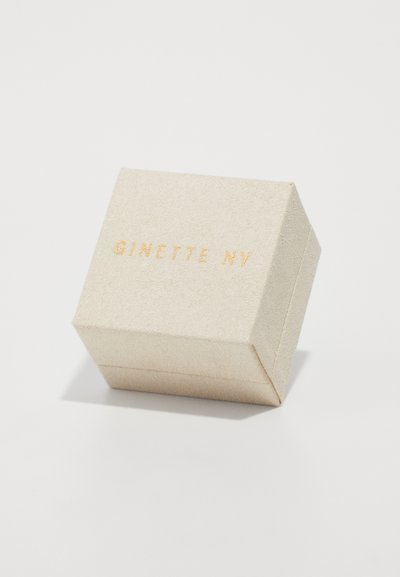 Caixa quadrada bege com superfície texturizada, logótipo em relevo dourado "GINETTE NY" na parte superior, apresentando linhas limpas e arestas nítidas.