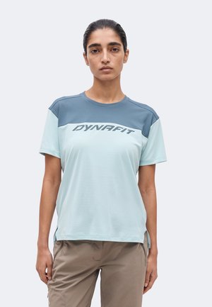 Γυναίκα που φοράει κοντομάνικη, δίχρωμη μπλε μπλούζα Dynafit και μπεζ παντελόνι, στέκεται μπροστά σε απλό φωτεινό φόντο.