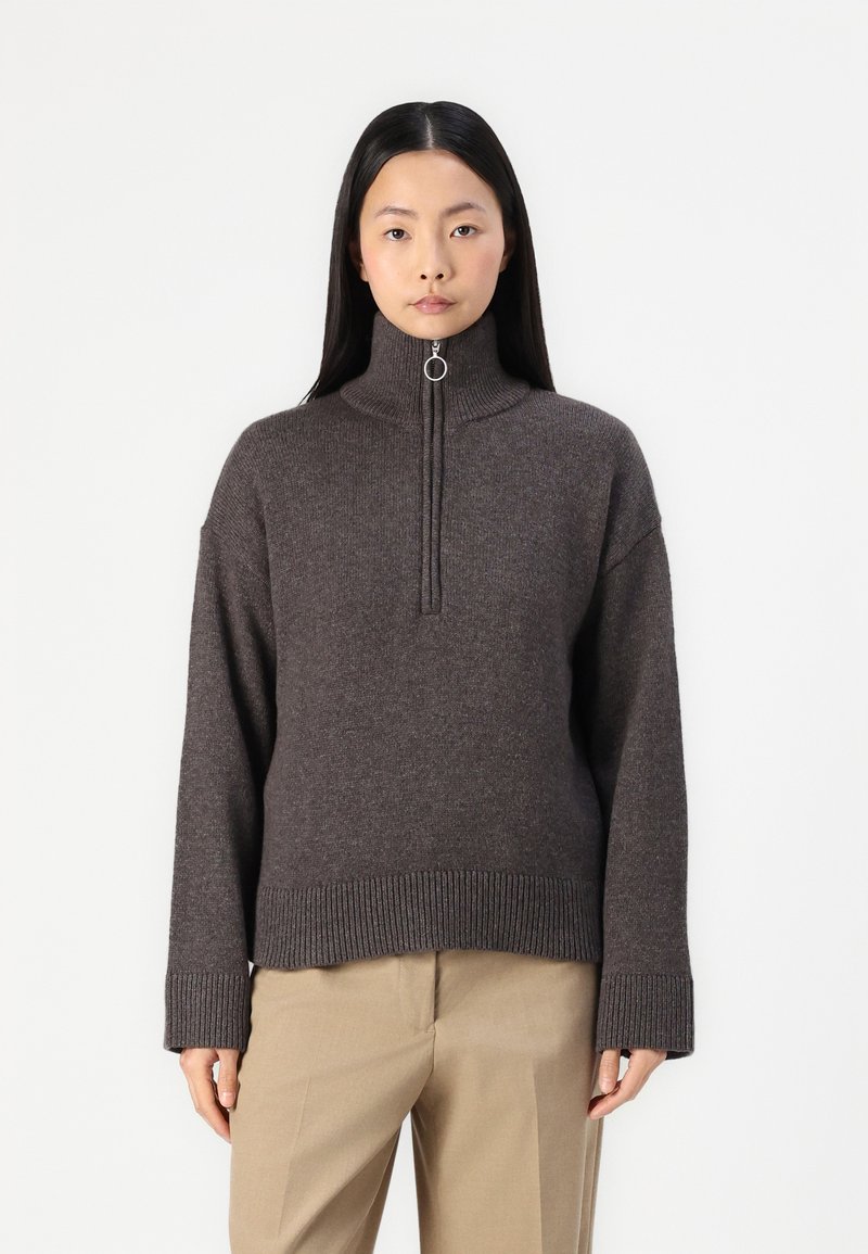 Brauner Strickpullover mit hohem Kragen, halber Reißverschluss und geripptem Saum. Weiche Textur mit legere Passform und überschnittenen Schultern.