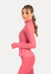 Top deportivo de manga larga rosa con cuello alto y agujeros para los pulgares, combinado con leggings a juego. Textura suave y diseño ajustado.